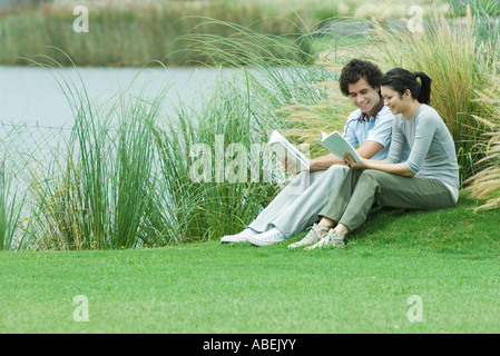 Young adult couple reading outdoors, sitting on grass by edge of lake Stock Photo