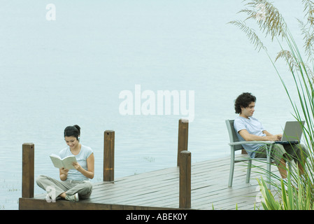 Couple sitting on dock, reading and using laptop Stock Photo