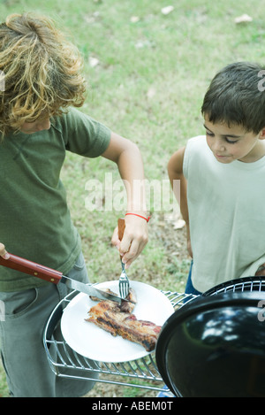 Two boys standing next to barbecue, one cutting up meat on plate Stock Photo