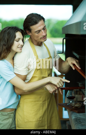 Man tending barbecue, while woman looks over his shoulder and points Stock Photo