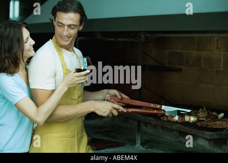 Man tending barbecue, while woman holds up glass of wine for him Stock Photo