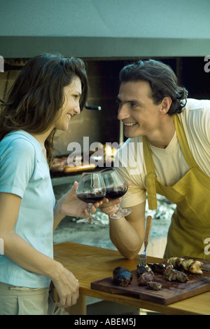 Couple standing in front of barbecue, clinking glasses of red wine and smiling at each other Stock Photo