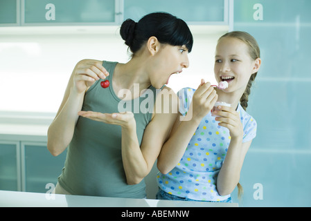 Girl holding ice cream dessert, next to woman holding cherry, woman leaning toward girl with mouth wide open Stock Photo