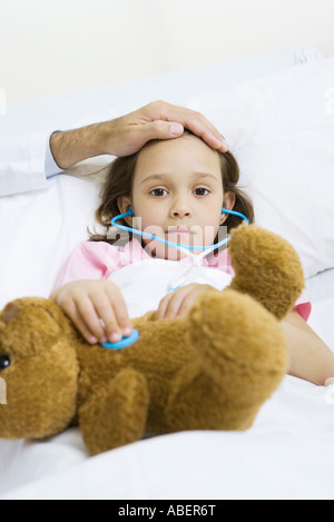 Child holding toy stethoscope to teddy bear, doctor feeling child's forehead Stock Photo