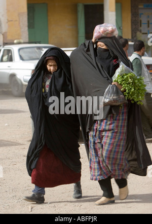 Two women in traditional black robes covering head carry vegetables street market Jorf in Tafilalt Morocco Stock Photo