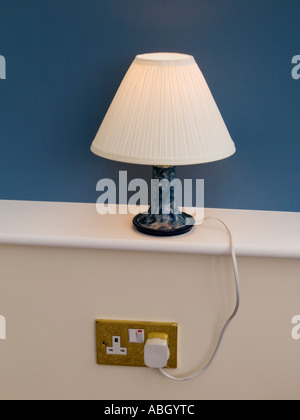 Table lamp lit up lamp shade on white windowsill with blue behind plugged into wall socket showing cable. England UK Britain Stock Photo