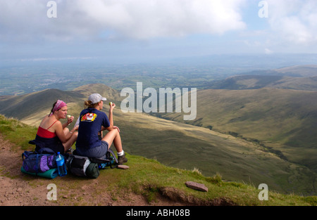 Two young women sit relaxing on the summit of Pen y Fan overlooking landscape of valleys Powys South Wales UK Stock Photo