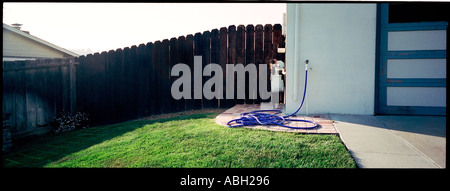 a suburban lawn with a wooden fence behind and a garage and driveway with a blue hose unevenly coiled on a small brick deck in B Stock Photo