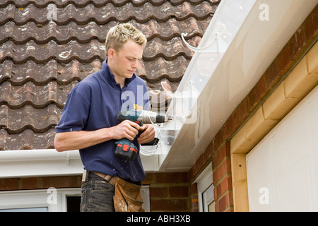 A builder using a cordless drill to install brackets to hold a gutter below a roof of a home. Stock Photo