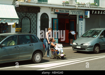 Lady manoeuvring wheelchair between parked cars in order to cross road Stock Photo