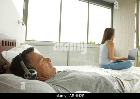 Couple in bedroom, man listening to music, woman using laptop, indoors Stock Photo