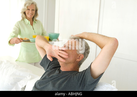Woman bringing breakfast to man yawning in bed Stock Photo