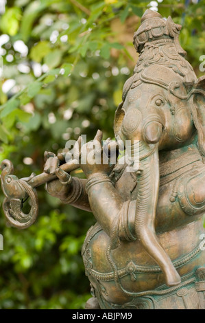 Intricate bronze sculpture of Hindu elephant god Ganesh or Ganesha playing a flute, set against a lush green garden background. Stock Photo