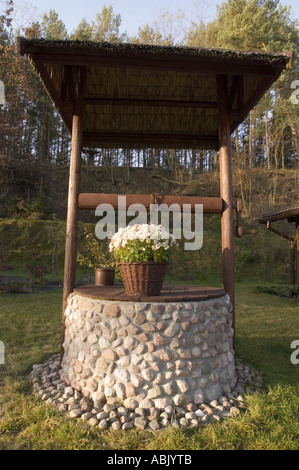 Pot with flowers standing on a decorative stone water well in a sunny garden with a forest background. Stock Photo