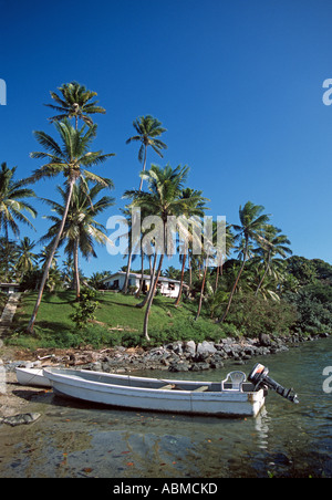 A small motorboat is docked on a rocky beach with palm trees a white house and a blus sky in the background Stock Photo