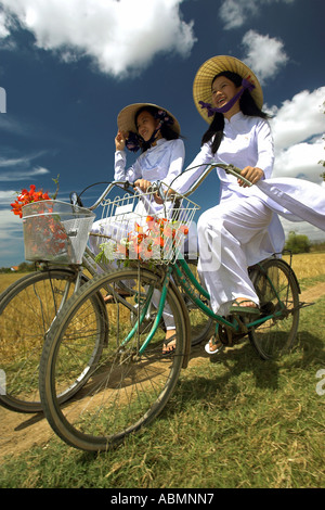 Two young women in conical hats and traditional costume ride bicycles through rice fields near Phan Thiet Vietnam Stock Photo