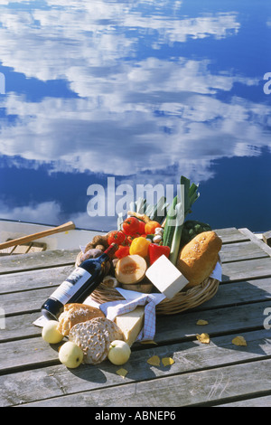 Picnic basket of fruits, bread and vegetables with bottle of red wine on lake pier Stock Photo
