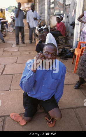 Disabled Ugandan businessman using his mobile phone crawls down street on his hands and knees Stock Photo