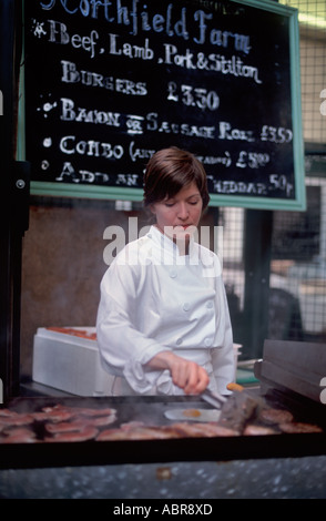 Young woman frying breakfasts on a smokey grill before farm shop sign promoting beef, lamb and pork, Borough Market, London Stock Photo