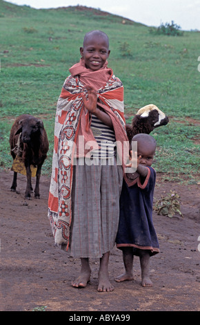 KENYA Masai Mara National Reserve Young Masai girl in traditional kanga cloth and her brother herd sheep near their village Stock Photo