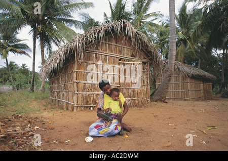 Woman and baby sitting outside a hut made from palm leaves used to store coconuts in Mozambique Stock Photo