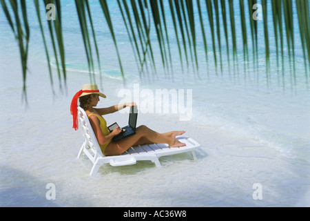 Woman sitting on sandy shore in beach chair with laptop during working holiday Stock Photo