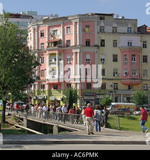 Tirana town centre street scene people crossing footbridge over stream ditch pastel coloured apartment block building beyond in Republic of Albania Stock Photo