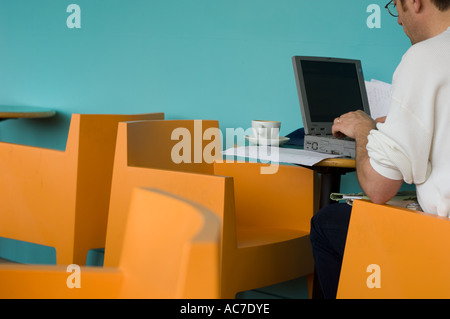 Man using a laptop computer in a trendy modern urban cafe, sitting on a orange plastic chair, UK Stock Photo