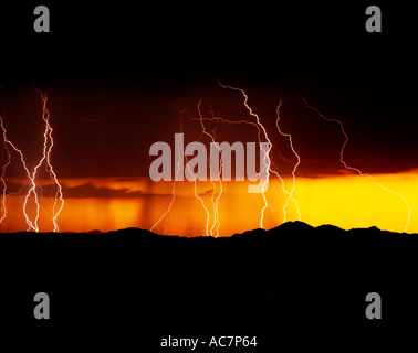 Lightning strikes during a vibrant orange and yellow sunset over mountains west of Tucson, AZ, USA. Rain falls behind the bolts. Stock Photo