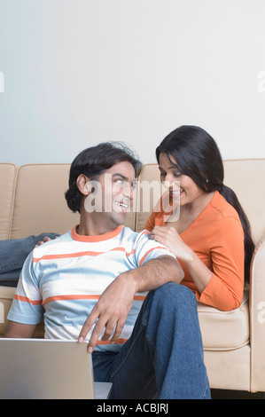 Young man sitting in front of a laptop with a young woman reclining on a couch behind him Stock Photo