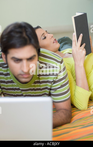 Mid adult man using a laptop with a young woman leaning on his back and reading a book Stock Photo