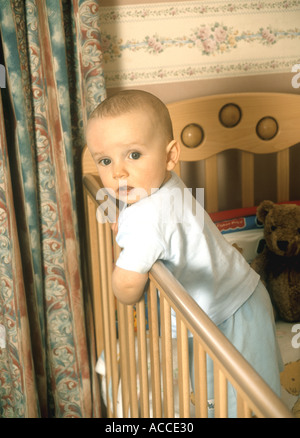 Baby boy leaning over a cot in his nursery Stock Photo