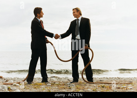 Two businessmen shaking hands on a beach holding a rope. Stock Photo