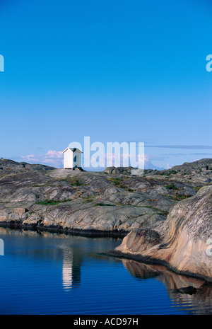 A small lighthouse on cliffs with calm reflecting water in the foreground and a blue sky in the background. Stock Photo