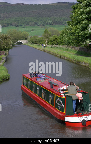Red & green narrowboat (hire boat) sailing on scenic rural Leeds Liverpool Canal on sunny day, 1 man standing by tiller - North Yorkshire, England, UK Stock Photo