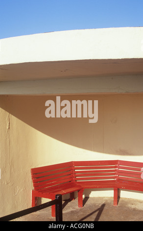 Detail of a red bench seat in a modernistic circular cream shelter casting an interesting curved shadow under a blue sky Stock Photo