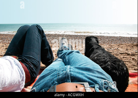A couple relaxing on the beach with their black labrador dog watch the sand and sea sitting on a rug Stock Photo