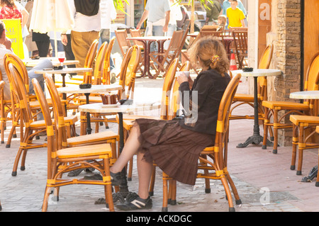 Cafe scene outdoor terrace in the old town, a young woman alone in big curly red hair drinking a cup of cafe au lait caffe latte, sitting on a wicker chair looking the other way, dressed in brown and a chequered skirt, running shoes Sanary Var Cote d’Azur France Stock Photo