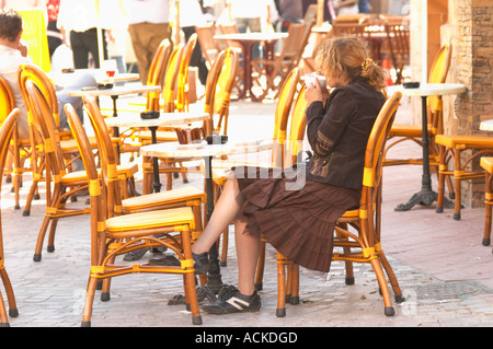 Cafe scene outdoor terrace in the old town, a young woman alone in big curly red hair drinking a cup of cafe au lait caffe latte, sitting on a wicker chair looking the other way, dressed in brown and a chequered skirt, running shoes Sanary Var Cote d’Azur France Stock Photo