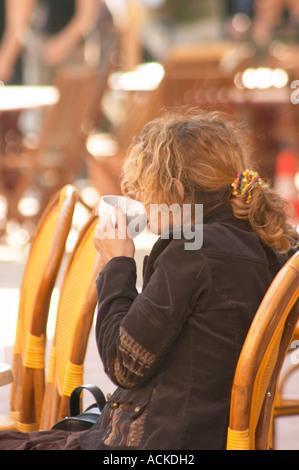 Cafe scene outdoor terrace in the old town, a young woman alone in big curly red hair drinking a cup of cafe au lait caffe latte, sitting on a wicker chair looking the other way, dressed in brown and a chequered skirt, running shoes Sanary Var Cote d’Azur France Stock Photo