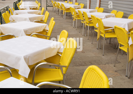 two rows of tables covered with white table cloths and surrounded by yellow wicker chairs in an outdoor cafe area in Venice Stock Photo
