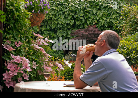 Male eating a large bread roll sandwich with a  "glass of Wine" on a table outside in a lush garden in summer Stock Photo