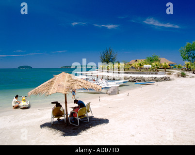 beach with white sand and two people under a rush/vegetation made umbrella sitting in the midday sun Stock Photo