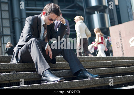 Young businessman puts his hand to his head in the City of London sitting on steps in front of the Lloyds building Stock Photo