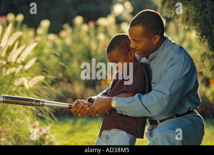 African American father teaches his son to swing a baseball bat in a park. Stock Photo