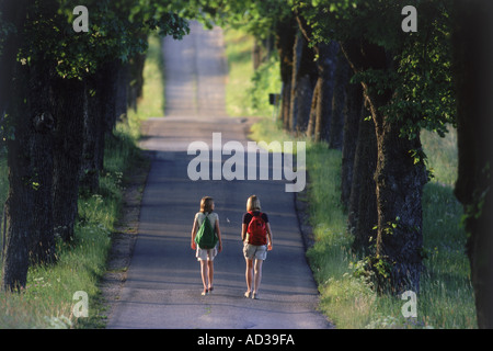 Two young girls with school backpacks walking on tree-lined country road in Sweden Stock Photo