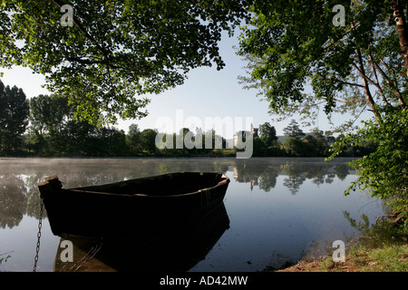 Wooden boat on flat calm water with a small chateau in the background beside the River Lot near Cahors in south west France Stock Photo