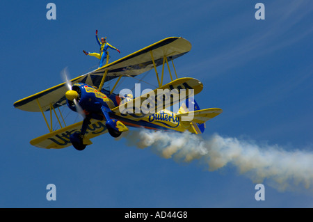 A bi-plane flying in the blue sky with a wing-walker on the top, and a smoke trail trailing behind. Stock Photo