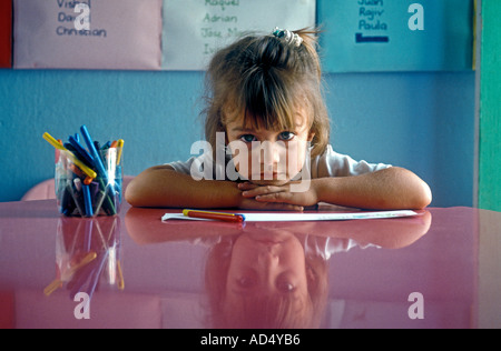 Infant Hispanic schoolgirl in kindergarten reception aged 3 to 4 years sitting pensive, in school classroom at a desk chin on hands with reflection Stock Photo