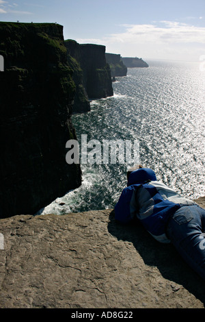 A young woman down on her stomach takes in the view of the Cliffs of Moher from the flat rock viewing place beyond the barrier Stock Photo